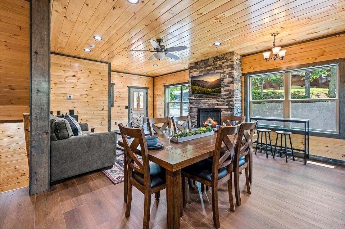 A wooden-paneled room with a dining table and chairs, a fireplace, and a large window.