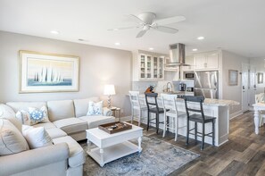 A modern living room with a sofa, coffee table, and a dining area with chairs and a kitchen in the background. - Pebble Beach D-206 3BR Oceanfront Condo (Emeral Isle)