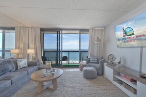 Un salon moderne doté d’une grande fenêtre donnant sur une plage, d’un téléviseur à écran plat et d’un coin salon confortable.