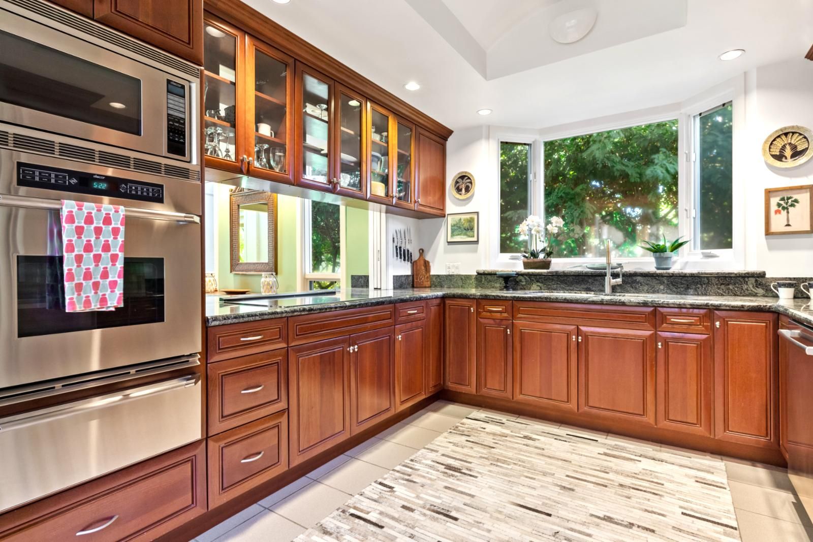 A modern kitchen with wooden cabinets, stainless steel appliances, and a large window providing a view of greenery.
