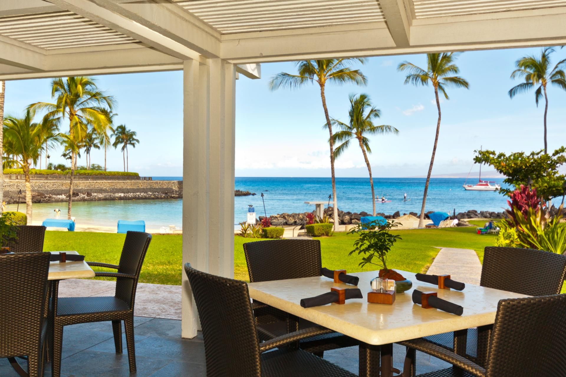 A covered outdoor seating area with wicker chairs and a table, overlooking a beach with palm trees and a boat in the distance.