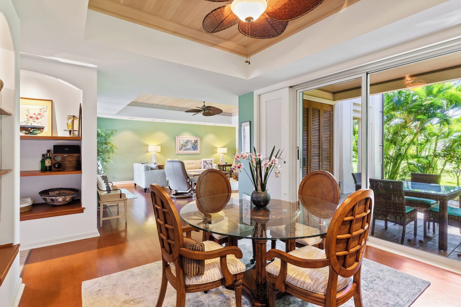 A dining area with a glass table and wooden chairs, a ceiling fan, and a view of a tropical garden through a sliding door.