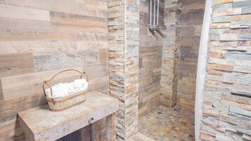 A shower area with stone walls, a wooden bench with a basket, and a grey rug.