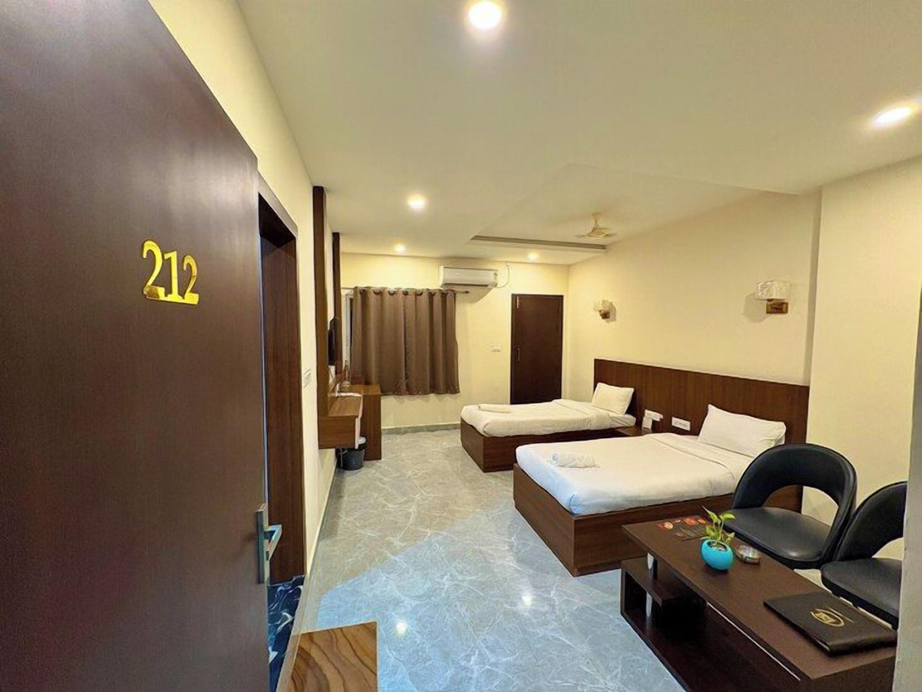 A hotel room with two beds, a desk, a chair, a small table with a plant, and a door marked with the number 212.