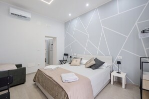 Moderni hotellihuone, jossa on sänky, tuoli, yöpöytä ja ilmastointilaite.