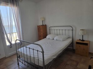 Ein Einzelbett mit Metallgestell, ein Nachttisch aus Holz mit einer Lampe und eine Holzkommode mit Lampe.