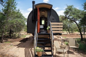 A small, dark-colored cabin with a staircase leading to a bed and a window. There are two wicker chairs outside. - Nomad Wagon With Private Hot Tub and Grill (Dripping Springs)