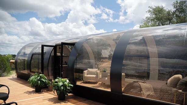 Una estructura transparente y alargada con terraza y zona de estar de madera, rodeada de follaje.