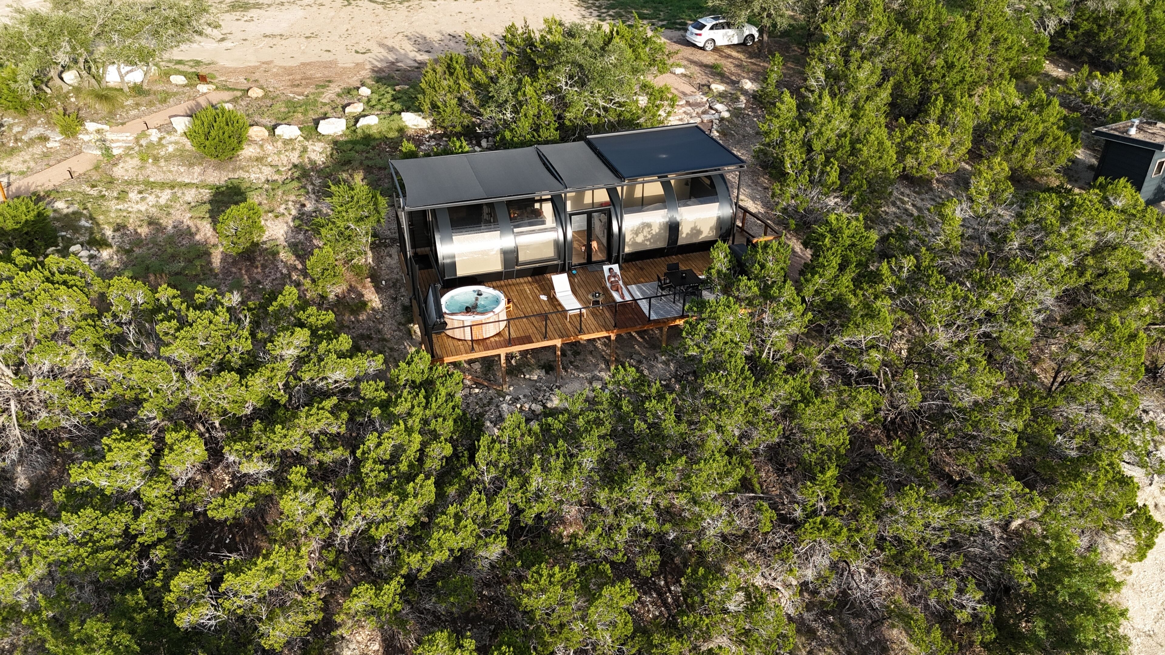 A modern two-story cabin with a deck, a hot tub, and a swimming pool surrounded by dense forest.