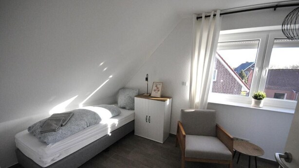Un dormitorio pequeño y moderno con cama alta, armario blanco, una silla y ventana con vistas a edificios.