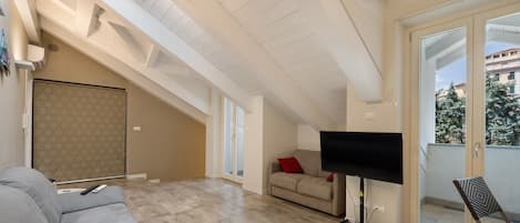 A spacious living area with a high ceiling, a sofa, a flat-screen TV, and a balcony with a view of buildings and trees.