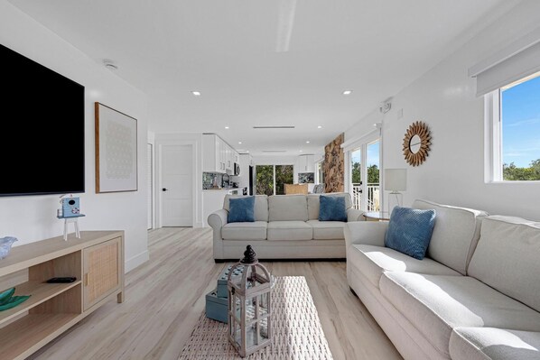 A modern living room with a large flat-screen TV, a sofa set, a coffee table, and a dining area with a kitchen in the background. - Hidden Keys Paradise-Where the Key Deer Roam-75’ Private Dock! Boaters Paradise! (Big Pine Key)
