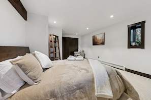 Ein Schlafzimmer mit Bett, Kissen, einer Hockerleiter, einem Sessel, einem Gemälde und einem Fenster.