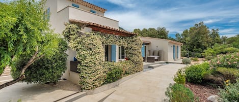 Una casa moderna con camino de entrada pavimentado, patio cubierto y abundante vegetación.