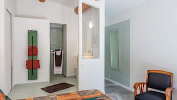 A room with a bed, a chair, a green towel rack, a bathroom with a shower, and a mirror.