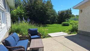 An outdoor patio with wicker furniture and a concrete table, surrounded by a grassy lawn and trees. - Snow's family friendly backyard oasis. YEG Airport. (Leduc)