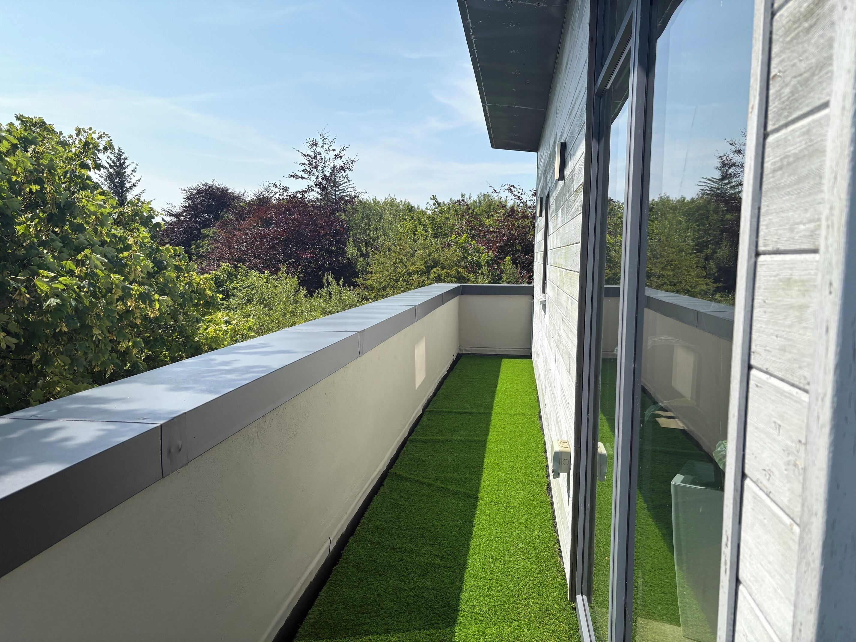 A modern balcony with a concrete edge, a glass door, and a view of greenery.