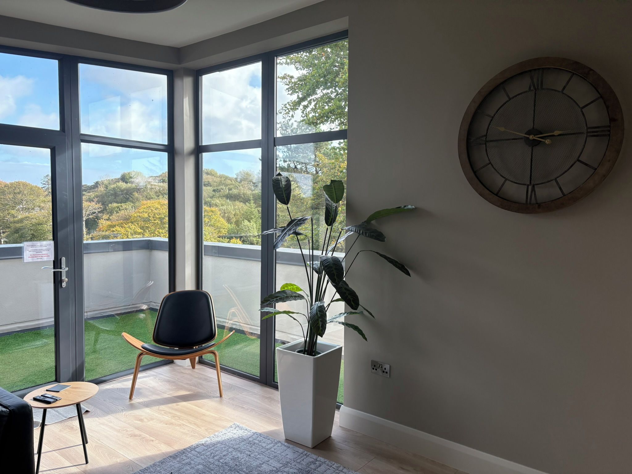 A modern room with large windows, a black chair, a potted plant, and a wall clock.