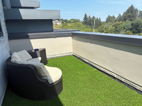 En takterrass med konstgräs, en flätad stol med kuddar och utsikt över ett bostadsområde.