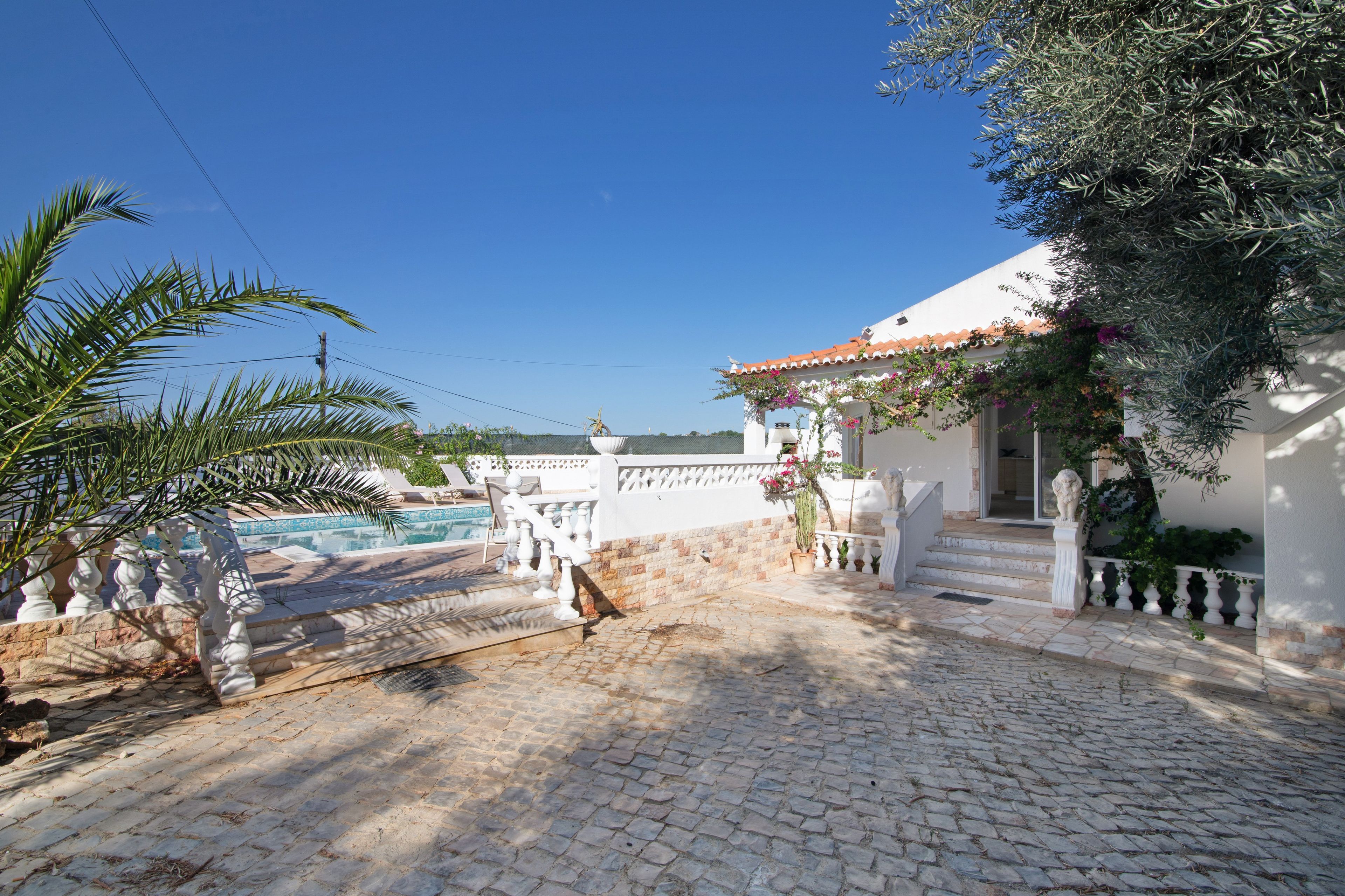 A cobblestone pathway leading to a white building with a tiled roof, a pool, and a palm tree.