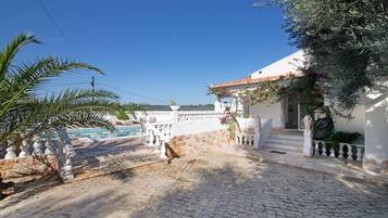A cobblestone pathway leading to a white building with a tiled roof, a pool, and a palm tree.