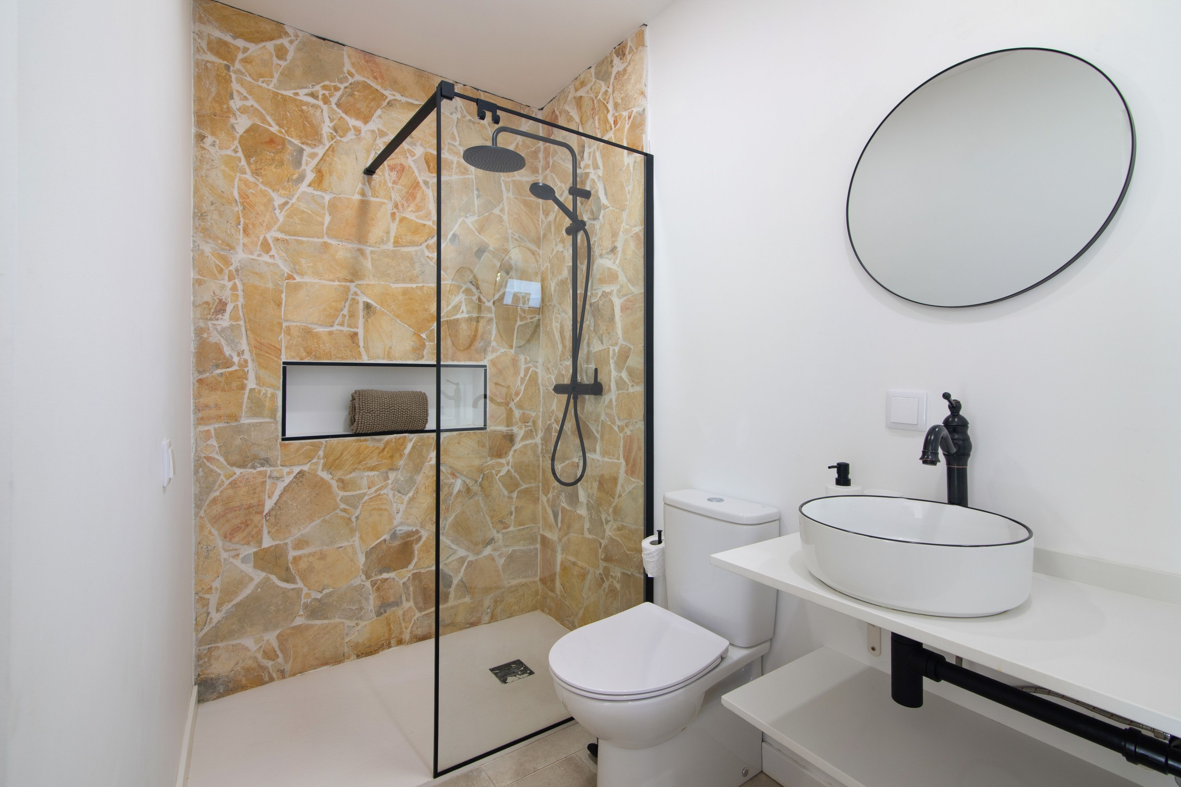 A bathroom with a stone wall, a glass shower enclosure, a toilet, a sink with a round basin, and a mirror.