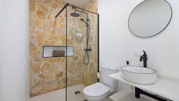 A bathroom with a stone wall, a glass shower enclosure, a toilet, a sink with a round basin, and a mirror.