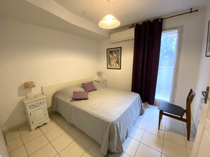 A bedroom with a bed, a nightstand, a chair, a window with curtains, and a painting on the wall. - "Le Capucine"—air conditioning, Wi-Fi, parking, downtown, harbor (La Ciotat)
