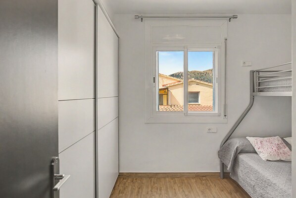 Ein kleiner, kompakter Raum mit einem Einzelbett, einem Fenster mit Blick auf ein Gebäude und Hügel sowie einem Kleiderschrank.