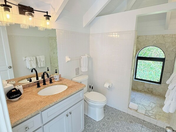 Ein Badezimmer mit doppeltem Waschbecken, Toilette, Dusche mit Fenster und Spiegel.