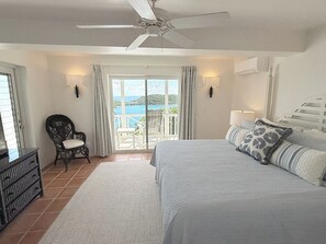 Ein Schlafzimmer mit Bett, einem Sessel, einem Deckenventilator und Meerblick durch eine Glastür.