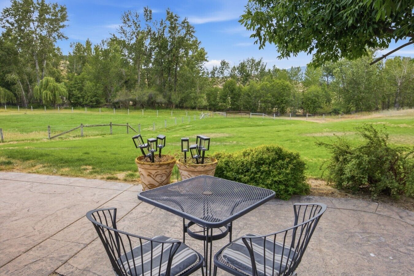 An outdoor patio with a metal table and chairs, surrounded by a grassy field and trees.