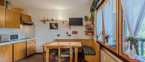 Una cocina compacta con armarios de madera, un microondas, una mesa de comedor con bancos y una ventana con cortinas.