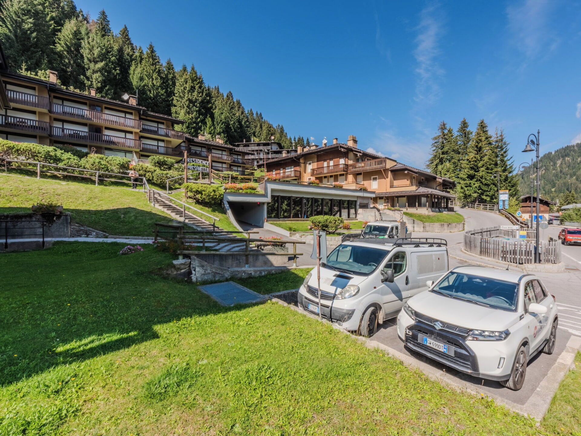 Un hotel con varias edificaciones, una zona de estacionamiento con múltiples autos y un paisaje montañoso de fondo.