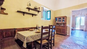 Un comedor con una mesa dispuesta para dos, sillas de madera, mantel estampado y una ventana con contraventanas azules.