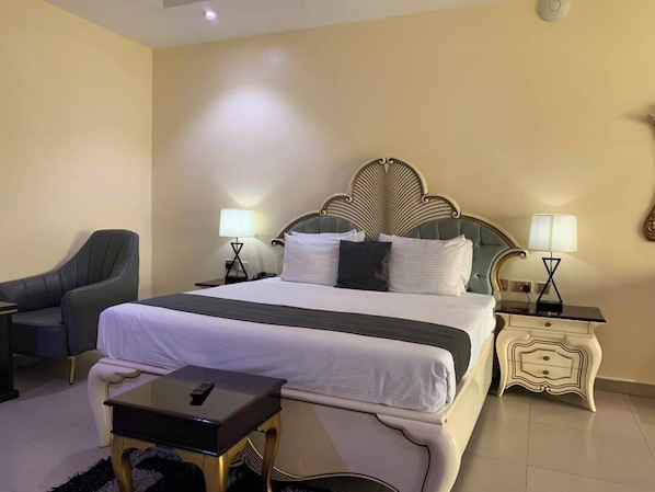 A hotel room with a large bed, two bedside tables with lamps, a chair, and a small table with a remote control. - Corporate Residence (Port Harcourt)