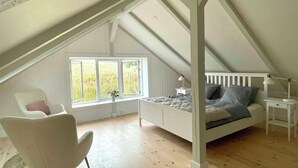 Ett sovrum med ett sluttande tak, en säng med kuddar, en stol, ett litet bord och ett fönster med utsikt över grönområden.