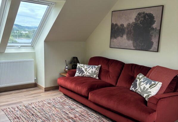 Ein rotes Sofa mit gemusterten Kissen, ein gerahmtes Landschaftsbild und Blick auf eine grüne Landschaft durch ein Oberlicht.