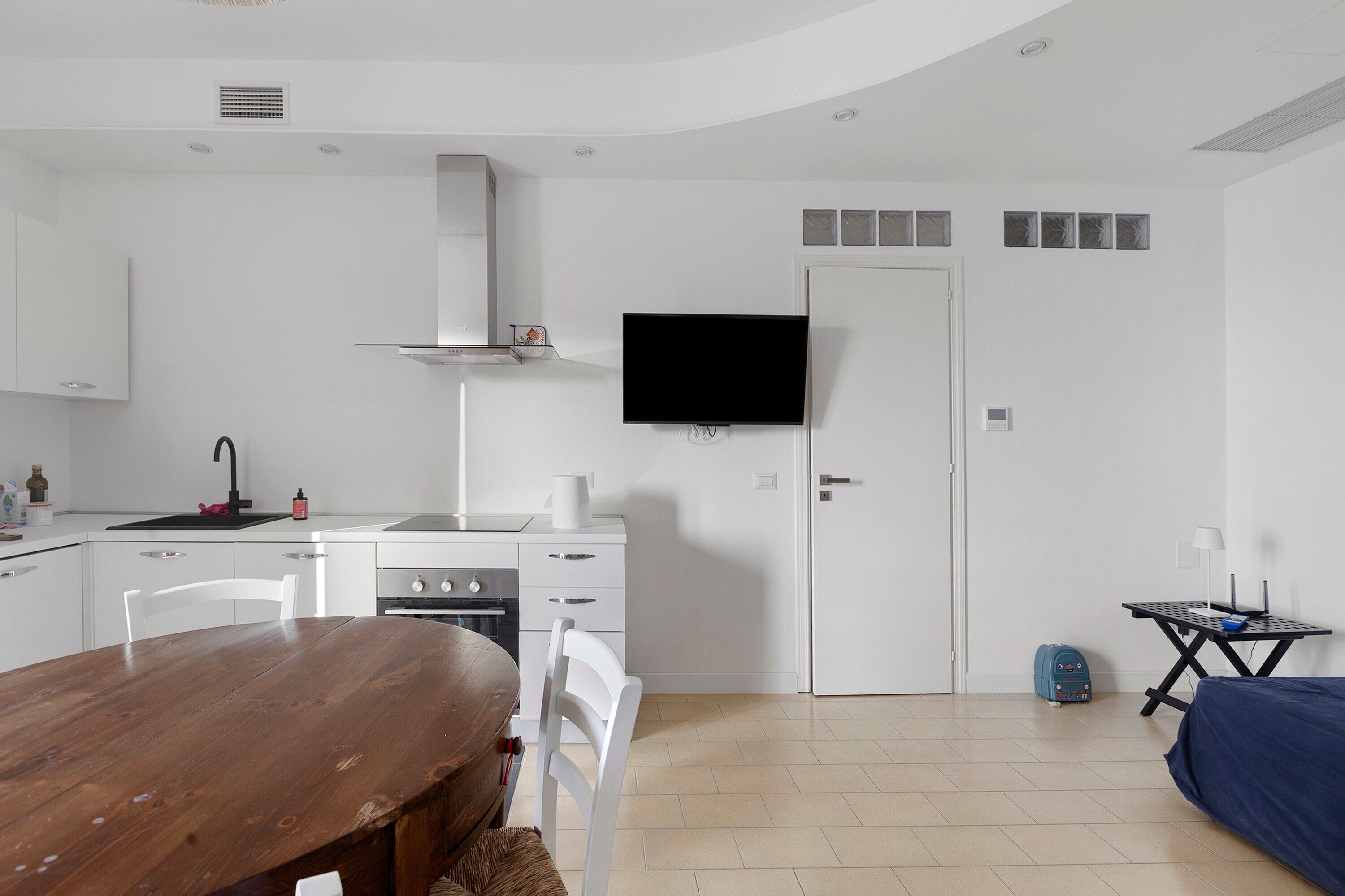 Uma cozinha compacta com armários brancos, uma mesa de jantar de madeira e uma televisão de tela plana presa à parede.
