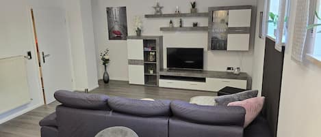 A modern living room with a grey sofa, a wooden dining table, and a flat-screen TV.