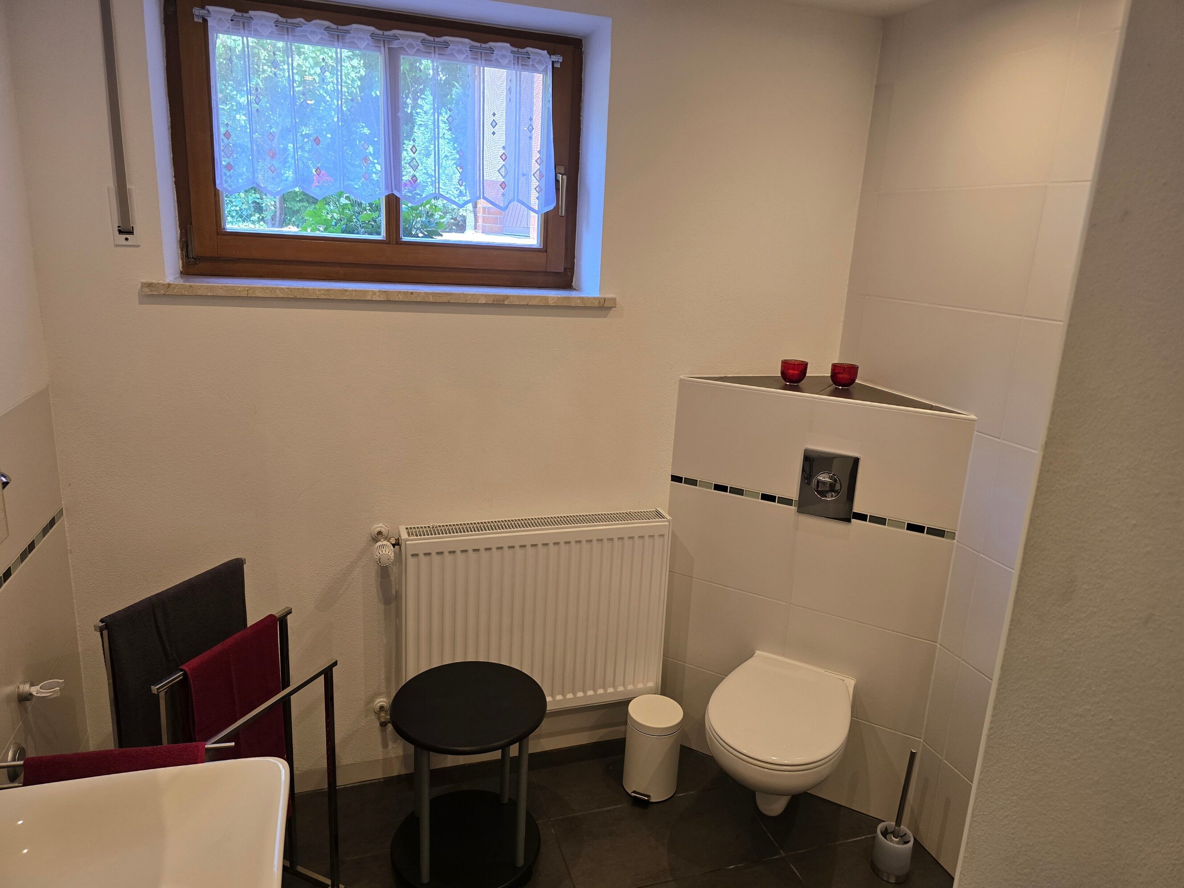 Une salle de bain comprenant des toilettes, un radiateur, une fenêtre avec un rideau et un porte-serviettes avec une serviette rouge.