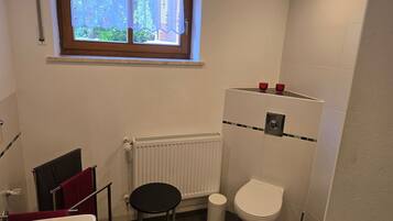 Une salle de bain comprenant des toilettes, un radiateur, une fenêtre avec un rideau et un porte-serviettes avec une serviette rouge.
