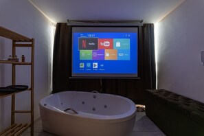 Quarto com uma tela grande exibindo interface de aplicativo de mídia, uma banheira, prateleira de madeira e um sofá.