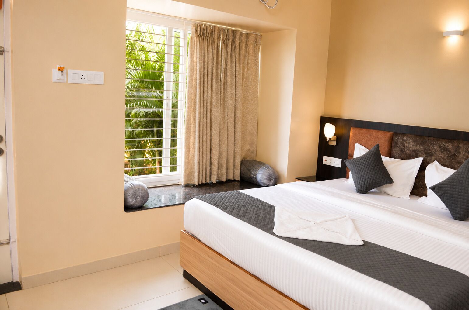 A hotel room with a bed, a window with curtains, a bedside table, and a wall-mounted light.