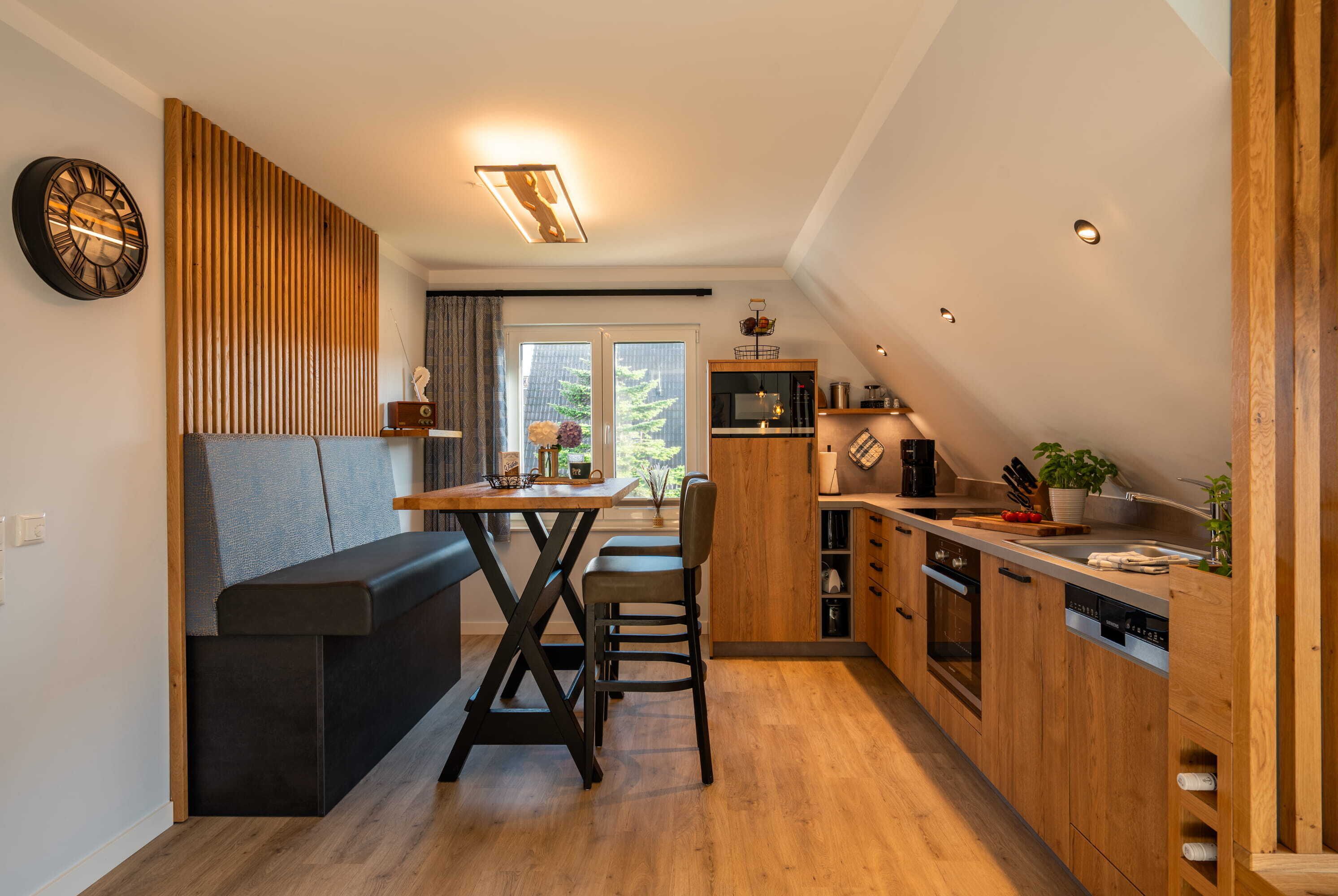 Een moderne keuken met houten kastjes, een zithoek met een bank en een groot raam dat voor natuurlijk licht zorgt.