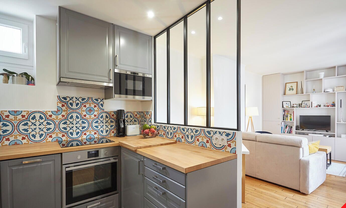 Una cocina moderna con gabinetes grises, horno empotrado y microondas. Cuenta con una encimera de madera y un salpicadero de azulejos con un estampado colorido.