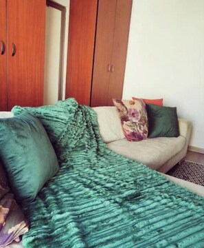 Ein Zimmer mit einem Bett, das mit einer grünen Decke bezogen ist, einem beigefarbenen Sofa mit einem Blumenkissen und Holzschränken.