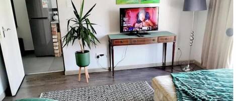 A room with a bed, a desk with a television, a plant, and a painting on the wall.
