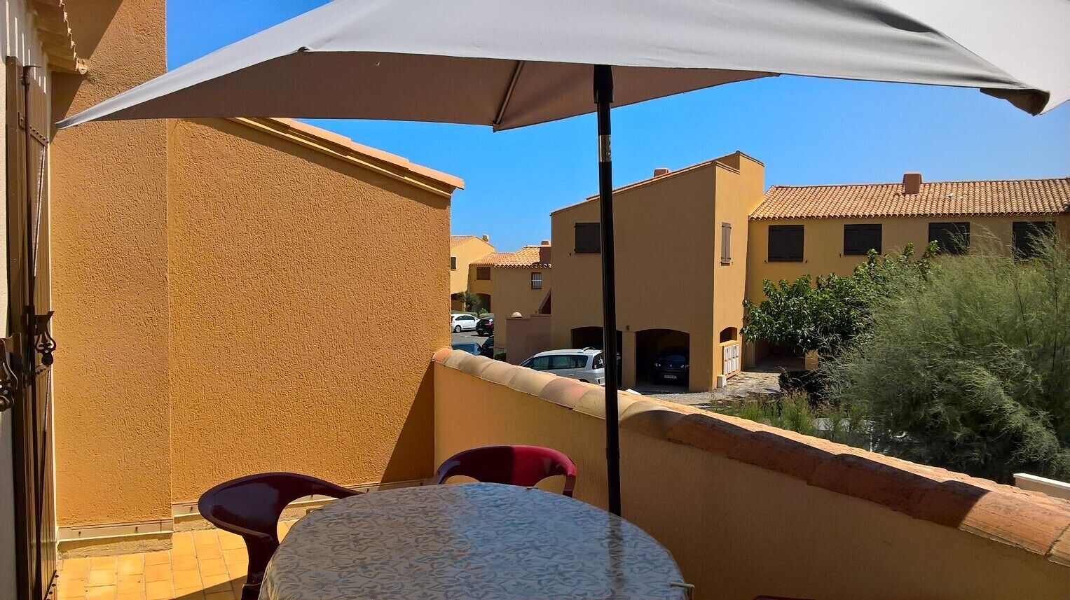 En balkon med et bord og stole, en stor parasol og udsigt til naboejengne.