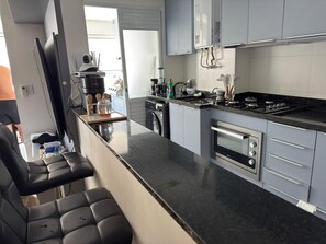 Une cuisine moderne équipée d’un four encastré, d’une cuisinière et d’une machine à café. On y trouve également un coin salon avec une chaise et une table.